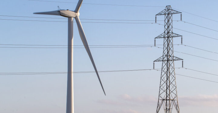 Un aerogenerador a la izquierda y una torre de alta tensión a la derecha. Ambos elementos están conectados por líneas eléctricas que se extienden a lo largo de la imagen. El cielo está despejado, con un tono azul claro, y la imagen sugiere un entorno relacionado con la producción y distribución de energía eléctrica.