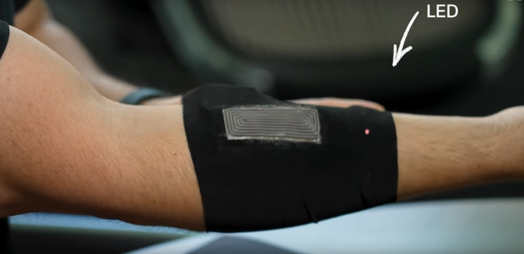 Un brazo con un dispositivo portátil flexible colocado sobre la parte superior del antebrazo. El dispositivo está diseñado para recolectar energía del calor corporal y convertirla en electricidad. En el extremo del dispositivo se observa un pequeño LED encendido, que indica que el calor del cuerpo está alimentando el sistema. El dispositivo tiene una apariencia delgada y está integrado en una prenda elástica, adaptándose al contorno del brazo.