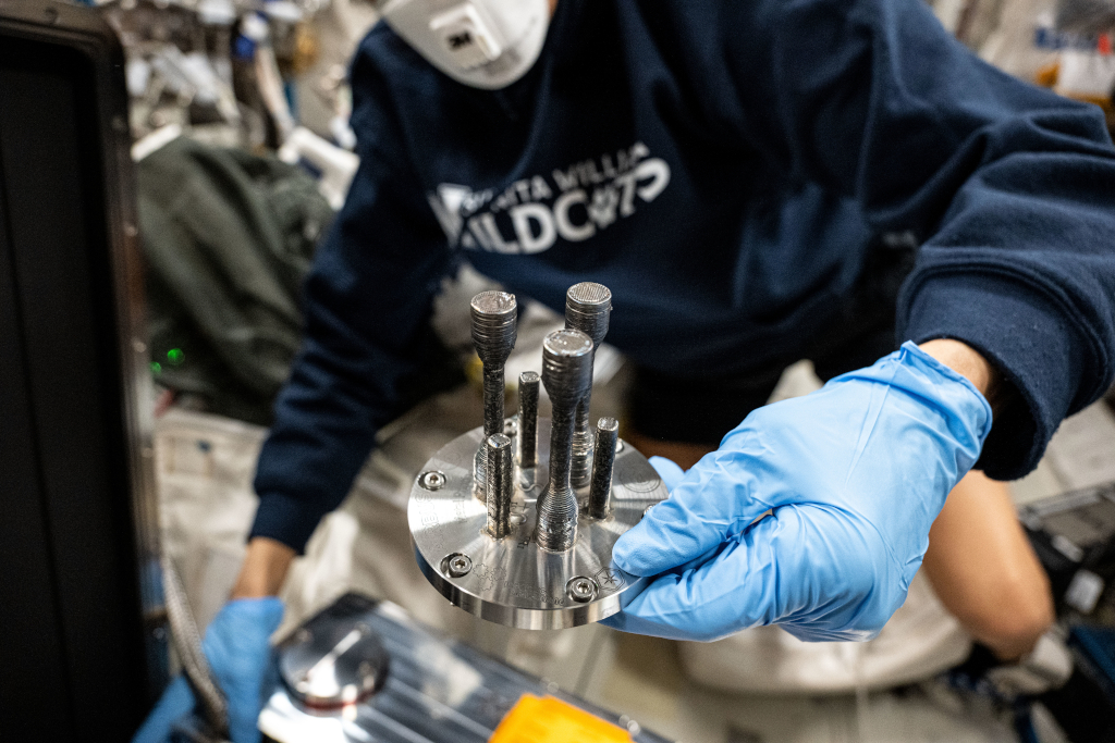 Un miembro de la tripulación de la Estación Espacial Internacional sostiene una placa metálica con varias piezas cilíndricas de acero inoxidable impresas en 3D. Las piezas fueron fabricadas utilizando la primera impresora 3D de metal en el espacio, parte de un experimento de la Agencia Espacial Europea en colaboración con Airbus. El tripulante lleva guantes y una mascarilla mientras manipula el equipo en un entorno controlado.