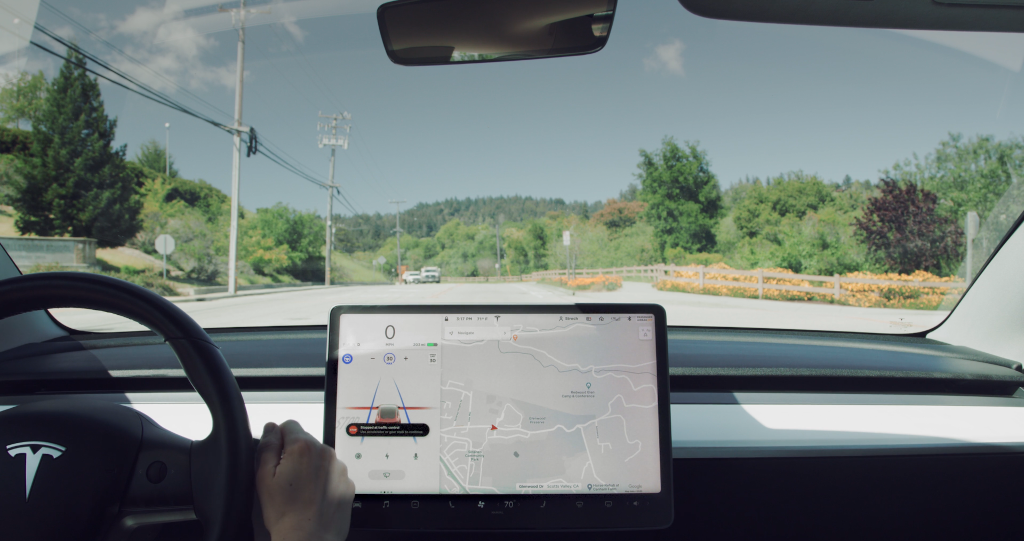 Vista desde el interior de un coche Tesla mientras circula por una carretera tranquila rodeada de vegetación. En el centro del salpicadero se observa la pantalla del sistema de navegación del vehículo, mostrando un mapa con indicaciones de conducción. El conductor sostiene el volante con una mano, y el sistema de "Full Self-Driving" parece activado. Al fondo, el cielo está despejado, y la carretera está despejada, sin otros vehículos visibles cerca.