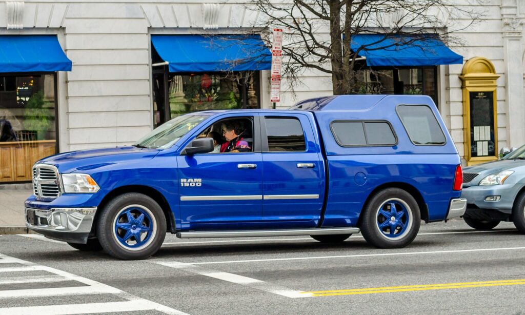 Una camioneta pickup Ram 1500 de color azul brillante circula por una calle urbana. El vehículo, de gran tamaño y con una cabina espaciosa, tiene llantas a juego con el color del cuerpo. Lleva una cubierta en la parte trasera, aumentando su volumen. En la acera se observan edificios comerciales con toldos azules y ventanas grandes. El conductor parece estar atento al tráfico, mientras que otro vehículo de menor tamaño se encuentra estacionado cerca. La escena refleja el contraste entre la camioneta y el entorno urbano.