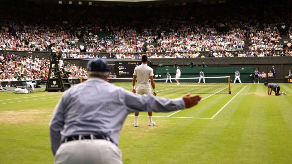 Un árbitro de tenis levanta el brazo y señala con firmeza durante un partido en una cancha de césped, mientras uno de los jugadores espera de espaldas. La imagen muestra una multitud de espectadores en las gradas, con el juez de silla sentado en su plataforma en el centro de la escena. Los recogepelotas y jueces de línea están atentos en el campo, en lo que parece ser un torneo de gran importancia, probablemente Wimbledon, por el ambiente y el césped característico.