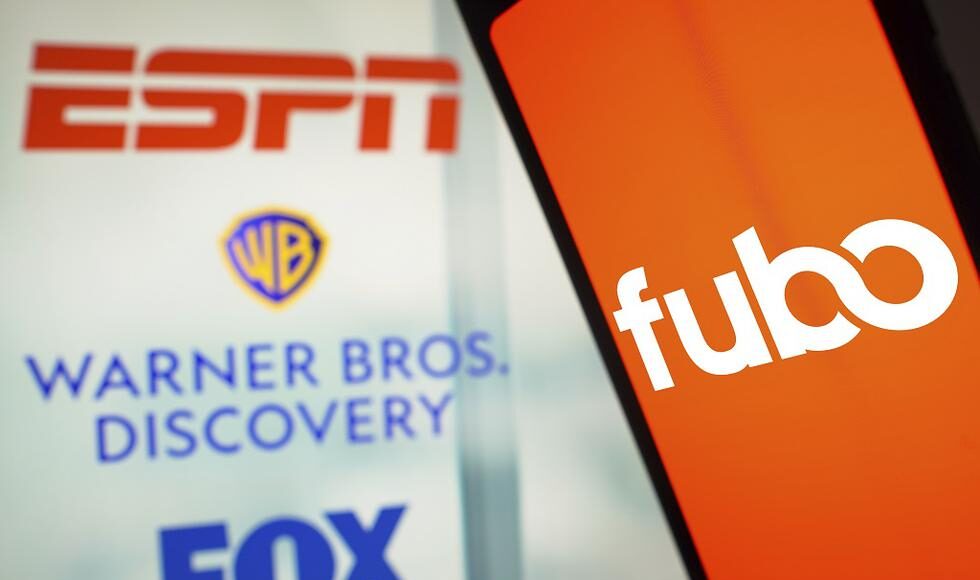 Logos de ESPN, Warner Bros. Discovery y Fox se muestran en un panel al fondo, mientras que en primer plano aparece la pantalla de un dispositivo móvil con el logotipo de Fubo en color blanco sobre un fondo naranja. La imagen representa la colaboración y acuerdos entre estas empresas en el contexto del streaming deportivo.