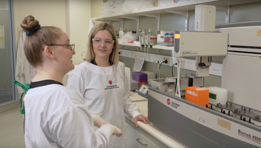 Dos investigadoras en un laboratorio científico, ambas usando batas blancas con el logo del QIMR Berghofer Medical Research Institute y gafas de seguridad. Una de ellas sostiene una conversación mientras están frente a un equipo automatizado de laboratorio de la marca Beckman Coulter, rodeado de tubos y cajas de reactivos. Al fondo, se observan estanterías con materiales y equipos de laboratorio.