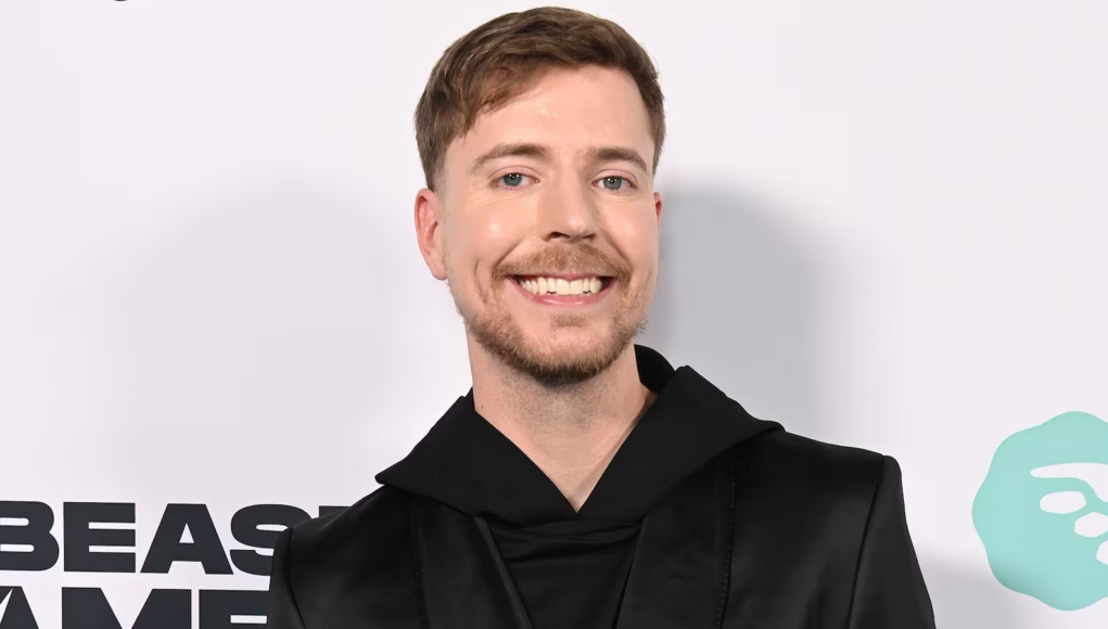 Una persona sonriente con cabello corto y barba ligera, vestida con una prenda negra de estilo moderno, posando frente a un fondo claro con logotipos parcialmente visibles.