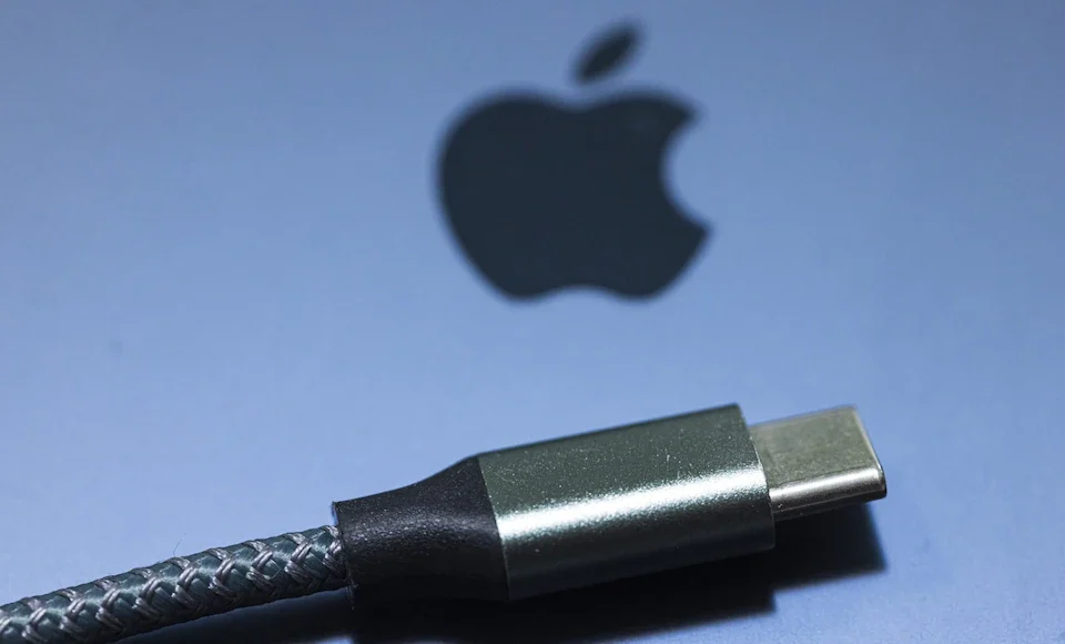 Cable USB-C con diseño trenzado en primer plano, colocado sobre una superficie gris con el logotipo de Apple difuminado al fondo.