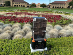 Dispositivo que utiliza el viento para generar amoníaco sin necesidad de energía externa Dispositivo experimental para la producción de amoníaco sostenible, instalado en un entorno al aire libre, rodeado de jardines florales y césped. El sistema incluye una malla recubierta con catalizadores montada sobre una base con tecnología portátil. Al fondo, se aprecian edificios universitarios y una avenida arbolada.