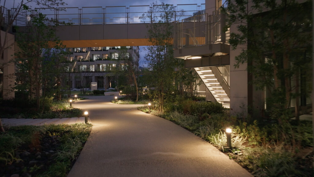 Sendero iluminado en Toyota Woven City rodeado de vegetación y diseño paisajístico moderno. Se aprecian edificios en el fondo con ventanas iluminadas, mientras un puente conecta estructuras, creando un ambiente armonioso entre naturaleza y urbanismo sostenible.