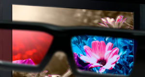 Unas gafas especiales en primer plano que muestran una vista dividida: el lado izquierdo de las lentes tiene un filtro rojo, mientras que el lado derecho muestra colores vibrantes y nítidos, como una flor rosa y azul. En el fondo, se observa una pantalla con flores que destacan por sus colores menos saturados fuera de las lentes.
