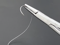 Puntos de sutura que generan electricidad al moverse para acelerar la curación Primer plano de una aguja curva con hilo de sutura sostenida por un portaagujas de metal, sobre un fondo negro.