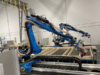 Empresas canadienses utilizan brazos robóticos portátiles e IA para ensamblar casas en un día Robots industriales azules en un almacén ensamblando paneles de madera para la construcción de viviendas. Los brazos robóticos, equipados con herramientas de precisión, trabajan sobre una estructura de madera, mostrando un proceso automatizado impulsado por inteligencia artificial. Al fondo, se observan elementos del entorno industrial, como cables colgantes y estanterías.