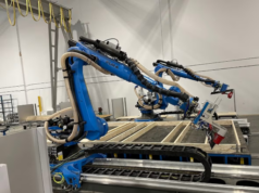 Empresas canadienses utilizan brazos robóticos portátiles e IA para ensamblar casas en un día Robots industriales azules en un almacén ensamblando paneles de madera para la construcción de viviendas. Los brazos robóticos, equipados con herramientas de precisión, trabajan sobre una estructura de madera, mostrando un proceso automatizado impulsado por inteligencia artificial. Al fondo, se observan elementos del entorno industrial, como cables colgantes y estanterías.