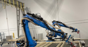 Robots industriales azules en un almacén ensamblando paneles de madera para la construcción de viviendas. Los brazos robóticos, equipados con herramientas de precisión, trabajan sobre una estructura de madera, mostrando un proceso automatizado impulsado por inteligencia artificial. Al fondo, se observan elementos del entorno industrial, como cables colgantes y estanterías.