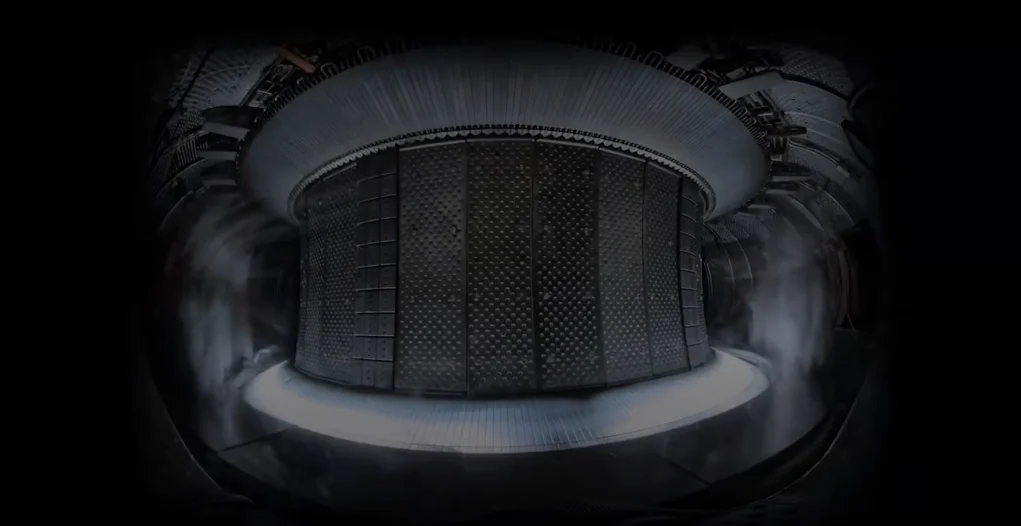 Interior de un reactor de fusión nuclear tipo Tokamak. Se observa una estructura cilíndrica metálica con paneles diseñados para resistir temperaturas extremas. En la parte inferior, un resplandor brillante indica la presencia de plasma de alta energía. Vapor o gas rodea la estructura, posiblemente como parte del sistema de enfriamiento o control del ambiente dentro del reactor. La iluminación tenue y el diseño industrial refuerzan la sensación de una instalación científica avanzada.