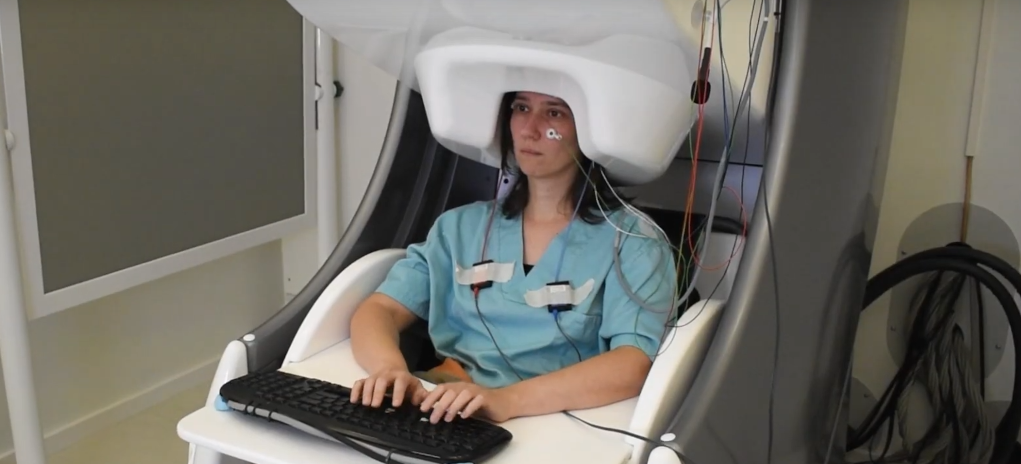 Mujer sentada frente a un teclado en un entorno de laboratorio, con un escáner de magnetoencefalografía (MEG) colocado sobre su cabeza. Lleva electrodos adheridos a su rostro y pecho, mientras participa en un estudio sobre la actividad cerebral durante la escritura.