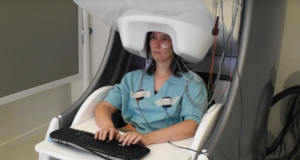 Mujer sentada frente a un teclado en un entorno de laboratorio, con un escáner de magnetoencefalografía (MEG) colocado sobre su cabeza. Lleva electrodos adheridos a su rostro y pecho, mientras participa en un estudio sobre la actividad cerebral durante la escritura.