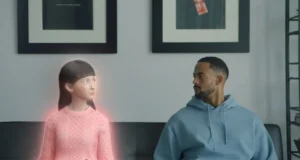 Un hombre con sudadera azul está sentado en un sofá, mirando a su lado, donde aparece una figura femenina digital con un brillo rosado. La figura parece ser un avatar o chatbot de inteligencia artificial con una apariencia estilizada y expresiva. El entorno es minimalista, con cuadros en la pared y una decoración moderna.