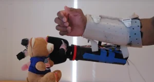 Prótesis de brazo biónico experimental utilizada por una persona. El brazo, construido con componentes mecánicos y electrónicos visibles, está sujeto con una estructura blanca y cintas de velcro. La prótesis se extiende hacia un osito de peluche con una camiseta azul y una bandera estadounidense en el pecho, simulando un intento de agarre. La escena se desarrolla sobre una mesa de madera con un fondo blanco.