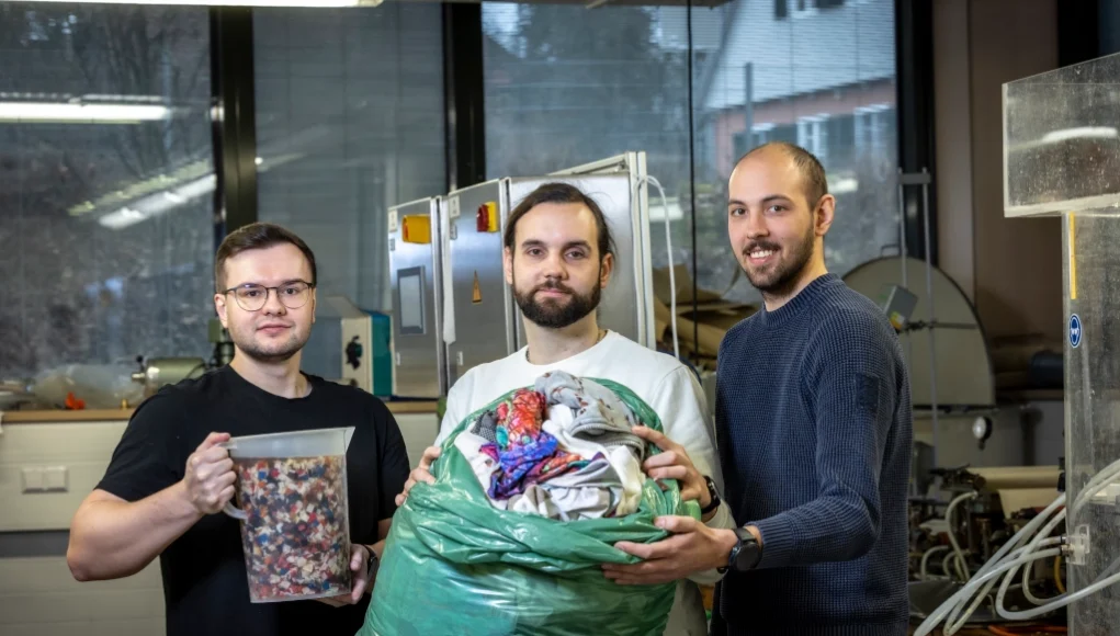 Tres hombres en un entorno de laboratorio sostienen materiales textiles reciclados: uno sujeta un recipiente con trozos de tela triturada, otro sostiene una gran bolsa verde llena de ropa usada, y detrás de ellos se ve maquinaria industrial y ventanas.