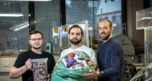 Tres hombres en un entorno de laboratorio sostienen materiales textiles reciclados: uno sujeta un recipiente con trozos de tela triturada, otro sostiene una gran bolsa verde llena de ropa usada, y detrás de ellos se ve maquinaria industrial y ventanas.