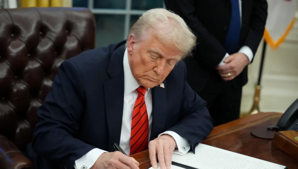 Un hombre de cabello rubio y piel clara, vestido con un traje oscuro, camisa blanca y corbata roja con rayas, está sentado en un escritorio de madera firmando un documento con un bolígrafo negro. Su expresión es seria y concentrada. En el fondo, se observa una persona de traje oscuro con las manos entrelazadas, así como una bandera y mobiliario característico de una oficina gubernamental.