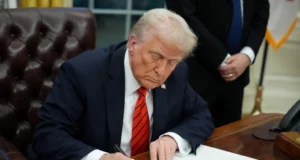 Un hombre de cabello rubio y piel clara, vestido con un traje oscuro, camisa blanca y corbata roja con rayas, está sentado en un escritorio de madera firmando un documento con un bolígrafo negro. Su expresión es seria y concentrada. En el fondo, se observa una persona de traje oscuro con las manos entrelazadas, así como una bandera y mobiliario característico de una oficina gubernamental.