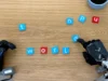 Google DeepMind lanza modelos de IA para un futuro más inteligente en robótica Dos manos robóticas manipulando pequeñas fichas con letras sobre una mesa de madera. Una de las manos está moviendo una ficha azul con la letra 'd'. En el centro de la imagen, las fichas forman la palabra 'world', con letras blancas sobre fondo azul y rojo. Otras fichas con letras dispersas están alrededor. En la esquina superior derecha, se ve parcialmente un objeto cilíndrico azul.