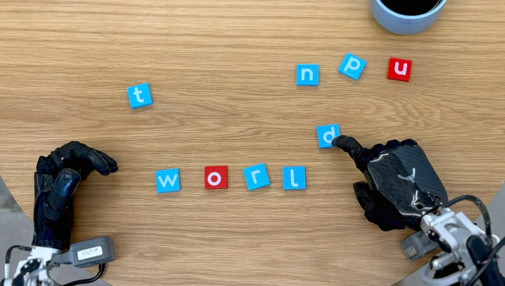 Dos manos robóticas manipulando pequeñas fichas con letras sobre una mesa de madera. Una de las manos está moviendo una ficha azul con la letra 'd'. En el centro de la imagen, las fichas forman la palabra 'world', con letras blancas sobre fondo azul y rojo. Otras fichas con letras dispersas están alrededor. En la esquina superior derecha, se ve parcialmente un objeto cilíndrico azul.