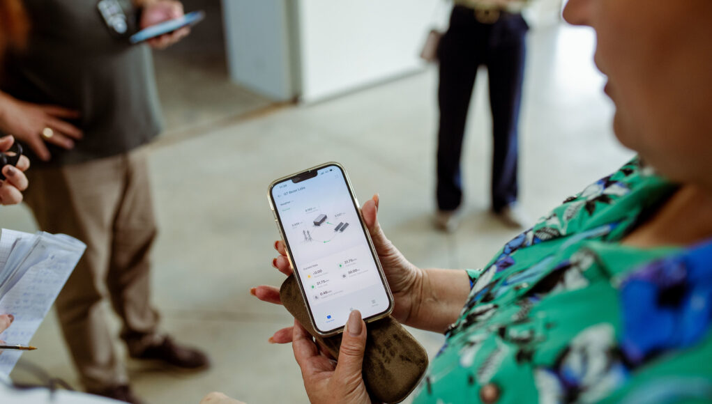 Primer plano de una mujer sosteniendo un teléfono inteligente que muestra datos técnicos en la pantalla, mientras otras personas toman apuntes y observan en segundo plano.