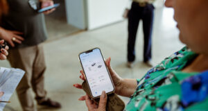Primer plano de una mujer sosteniendo un teléfono inteligente que muestra datos técnicos en la pantalla, mientras otras personas toman apuntes y observan en segundo plano.