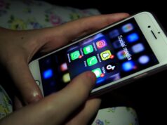 Persona sosteniendo un iPhone blanco con la pantalla encendida, mostrando una carpeta titulada "Social media" que contiene varias aplicaciones populares como Instagram, Snapchat, TikTok, WhatsApp, FaceTime y Phone. El entorno está oscuro y la luz de la pantalla ilumina ligeramente las manos del usuario.
