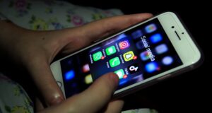 Persona sosteniendo un iPhone blanco con la pantalla encendida, mostrando una carpeta titulada "Social media" que contiene varias aplicaciones populares como Instagram, Snapchat, TikTok, WhatsApp, FaceTime y Phone. El entorno está oscuro y la luz de la pantalla ilumina ligeramente las manos del usuario.