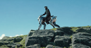 Persona montando un vehículo robótico cuadrúpedo de aspecto futurista sobre una formación rocosa al aire libre. El robot, con diseño metálico y articulaciones visibles, simula la forma de un animal de cuatro patas, mientras el piloto lleva casco y vestimenta de protección bajo un cielo despejado.