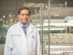 Un chicle antiviral para reducir la transmisión del virus de la gripe y el herpes simple Retrato de un científico sonriente, con bata blanca que lleva bordado "Henry Daniell, Ph.D." y el logotipo de Penn Dental Medicine. Está de pie en un laboratorio con estanterías metálicas iluminadas que contienen recipientes con plantas en cultivo.