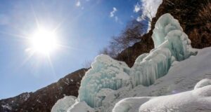 Formaciones de hielo y nieve en una ladera montañosa bajo un cielo despejado, con el sol brillando intensamente y árboles sin hojas al fondo, creando un contraste entre el clima frío y la luz solar.