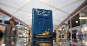 Un ejemplar de “The Great Gatsby” de F. Scott Fitzgerald, con su icónica portada azul mostrando un rostro con ojos y labios rojos sobre luces de la ciudad, se exhibe sobre una superficie de cristal en un espacio tipo feria o exposición, mientras se distingue la figura desenfocada de una persona al fondo.