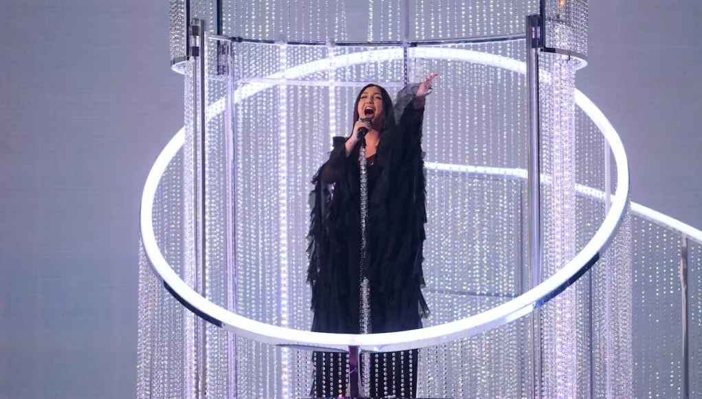 Mujer cantando con un micrófono en una plataforma elevada rodeada de estructuras circulares y decoraciones brillantes, bajo una iluminación escénica intensa.