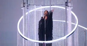 Mujer cantando con un micrófono en una plataforma elevada rodeada de estructuras circulares y decoraciones brillantes, bajo una iluminación escénica intensa.