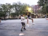 Dos hombres jugando kickball en una cancha urbana al atardecer; uno intenta alcanzar o bloquear al otro mientras sostiene una pelota verde, en una acción intensa y dinámica. Al fondo se observan más personas, árboles y edificios residenciales.