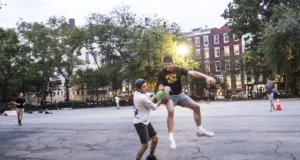 Dos hombres jugando kickball en una cancha urbana al atardecer; uno intenta alcanzar o bloquear al otro mientras sostiene una pelota verde, en una acción intensa y dinámica. Al fondo se observan más personas, árboles y edificios residenciales.