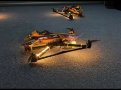¡Histórico! Un dron con IA vence por primera vez a campeones humanos en carreras Fotografía de varios drones de carreras iluminados, alineados sobre una superficie de alfombra gris. Los drones tienen un diseño aerodinámico con carenado en tonos morado, naranja y azul, y luces LED cálidas en sus brazos. Se observan logotipos de patrocinadores como "TU Delft", "DCL", "AWS" y "DJI". La imagen transmite una atmósfera tecnológica y competitiva, posiblemente en preparación para una carrera en interiores.