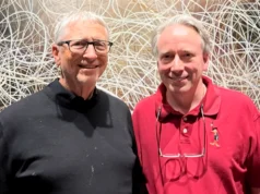 Bill Gates y Linus Torvalds se reúnen por primera vez en una cena histórica Dos hombres posan sonrientes frente a una pared con un fondo de líneas entrelazadas; uno lleva gafas y un suéter negro, y el otro una camiseta tipo polo roja con un dibujo bordado de un personaje animado.