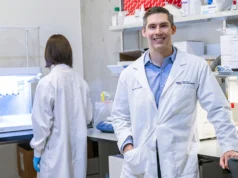 Investigadores desarrollan espermatozoides artificiales con IA y bioimpresión 3D para tratar la infertilidad masculina Dos investigadores en un laboratorio moderno; uno de ellos, un hombre con bata blanca y camisa azul, sonríe mientras mira a cámara, mientras que otra persona trabaja de espaldas frente a un equipo científico.