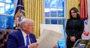 Un hombre de cabello rubio, vestido con traje azul, sostiene un documento oficial sentado en un escritorio, mientras una mujer vestida de negro lo observa de pie con una expresión de respeto. Están en una oficina con cortinas doradas, banderas oficiales y teléfonos sobre el escritorio.