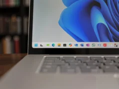 Microsoft confirma que Windows ha perdido hasta 400 millones de usuarios desde el lanzamiento de Windows 11 Primer plano del borde inferior de la pantalla de un portátil con Windows 11, mostrando el menú de aplicaciones ancladas en la barra de tareas. En el fondo, se aprecian estanterías desenfocadas llenas de libros.