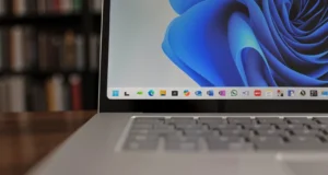 Primer plano del borde inferior de la pantalla de un portátil con Windows 11, mostrando el menú de aplicaciones ancladas en la barra de tareas. En el fondo, se aprecian estanterías desenfocadas llenas de libros.
