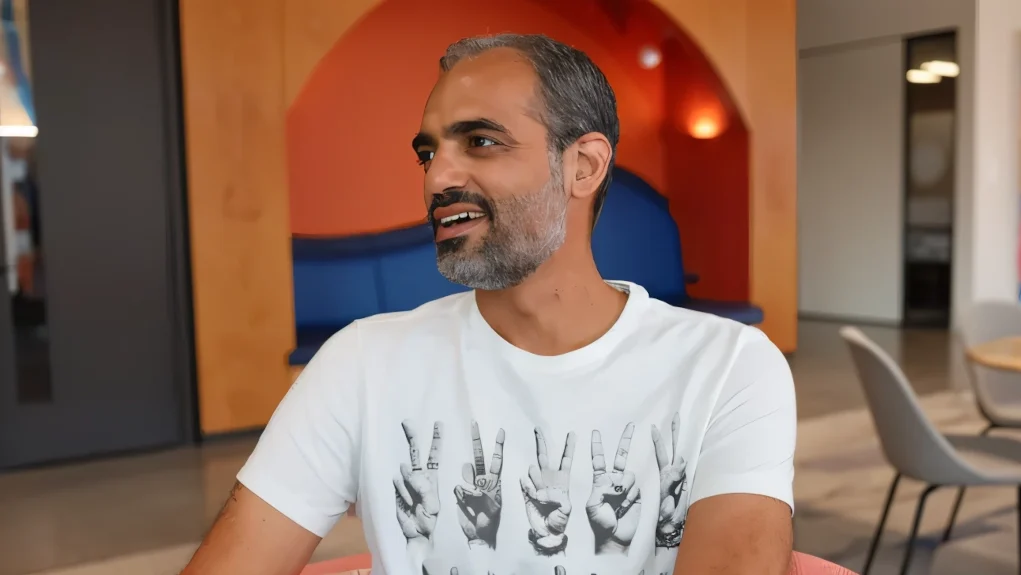 Hombre sonriente con barba entrecana y camiseta blanca con diseño de manos haciendo gestos, sentado en un espacio moderno de oficina con mobiliario contemporáneo y colores cálidos al fondo.
