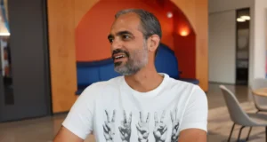 Hombre sonriente con barba entrecana y camiseta blanca con diseño de manos haciendo gestos, sentado en un espacio moderno de oficina con mobiliario contemporáneo y colores cálidos al fondo.