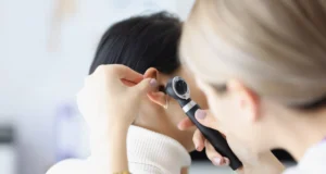 Una profesional médica examina el oído de una paciente usando un otoscopio. La paciente, de espaldas, tiene el cabello oscuro recogido y lleva un suéter blanco. La médica sostiene el instrumento con cuidado, enfocada en la revisión.