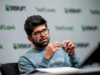 Hombre joven con gafas y barba hablando en un evento tecnológico, con un micrófono de diadema y gesticulando con las manos. Lleva una sudadera azul con la palabra "Alexity" impresa, y al fondo se distingue el logo de "Disrupt" repetido en el panel del escenario.